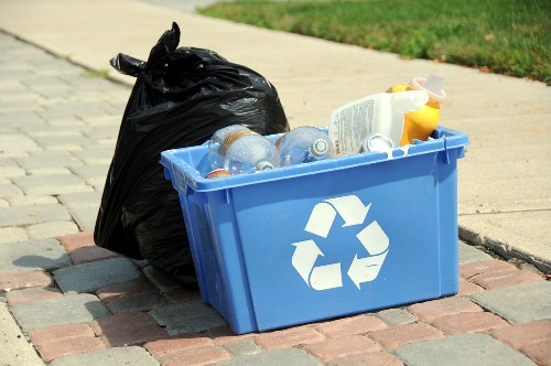 A blue recycling bin with a white recycling symbol on the side, filled with several transparent plastic bottles and some cardboard packaging, is positioned on a paved pavement near a curb. Behind the bin, there is a large black plastic rubbish bag that appears to be filled with waste. The background shows a grassy area and a section of a concrete pavement, indicating an outdoor setting in a suburban or residential area. The scene suggests waste collection or disposal activities typical in urban areas such as Euston or nearby locations, with a focus on recyclable materials and general refuse. The lighting indicates daylight, highlighting the textures of the plastic, cardboard, and paving stones, consistent with a typical outdoor waste management context involving rubbish removal services.