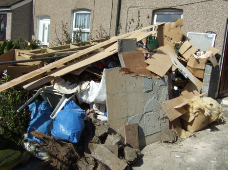 A pile of mixed waste materials situated on a paved area outside a residential building, featuring broken concrete blocks, pieces of drywall with textured grey surfaces, and various cardboard boxes, some flattened and torn, in different shades of brown. Among the debris, there are wooden planks, one partially leaning against the wall, and a blue plastic bag containing unidentified rubbish. The background shows the front of a beige pebble-dashed house with white-framed windows and small garden plants, indicating a typical outdoor space in Euston. The scene appears to be part of a rubbish removal process, with the waste accumulated on the driveway or pavement area. This image illustrates the type of waste managed by Garden Clearance Euston, a company specializing in outdoor rubbish clearance services in the local area.