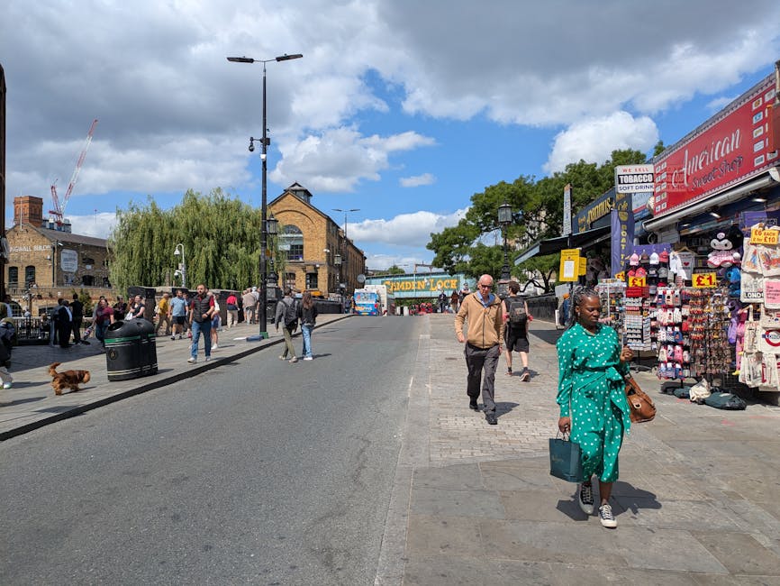 A busy urban street scene during daytime with pedestrians walking along the pavement and loading area. In the foreground, a woman wearing a bright turquoise polka-dot dress and white sneakers holds a 