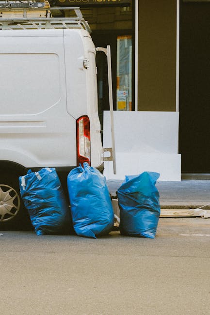 A narrow urban alleyway lined with graffiti-covered walls on both sides, cluttered with an assortment of discarded waste materials. In the foreground, a large, weathered black garbage bag, partially t